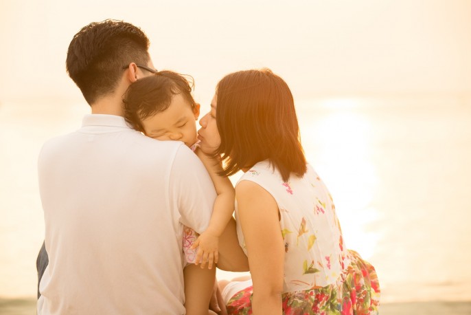 Portrait of young Asian family seated on beach outdoor vacation, during summer sunset, natural sunlight with flare.