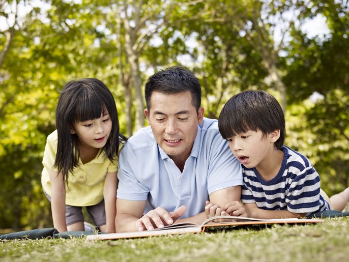 asian father and children lying on grass reading a book together in a park.